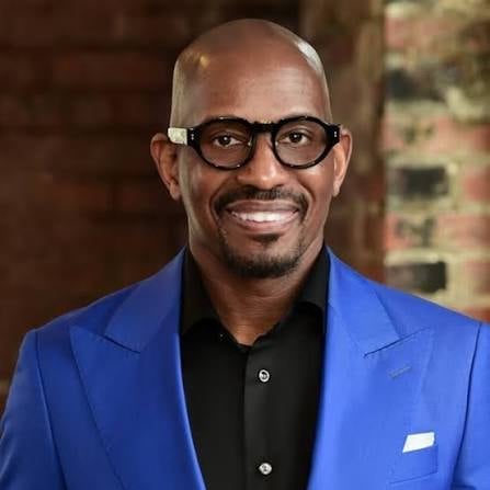 Professional headshot of a man wearing a bright blue blazer and black shirt, smiling at the camera with glasses