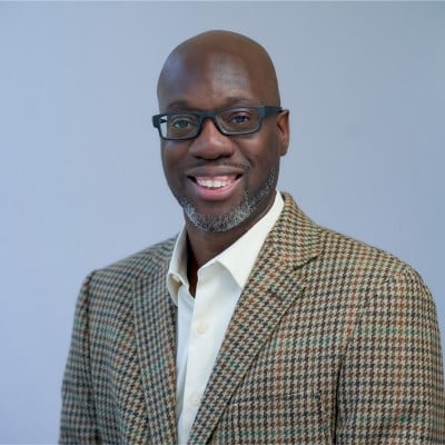 Professional headshot of a smiling man wearing glasses, a patterned blazer, and white shirt against a light blue background