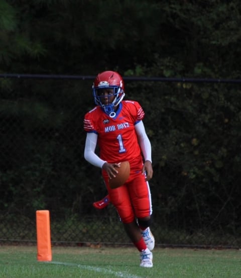 Young football player in red Mob Bots uniform with number 1 running with ball near orange end zone marker