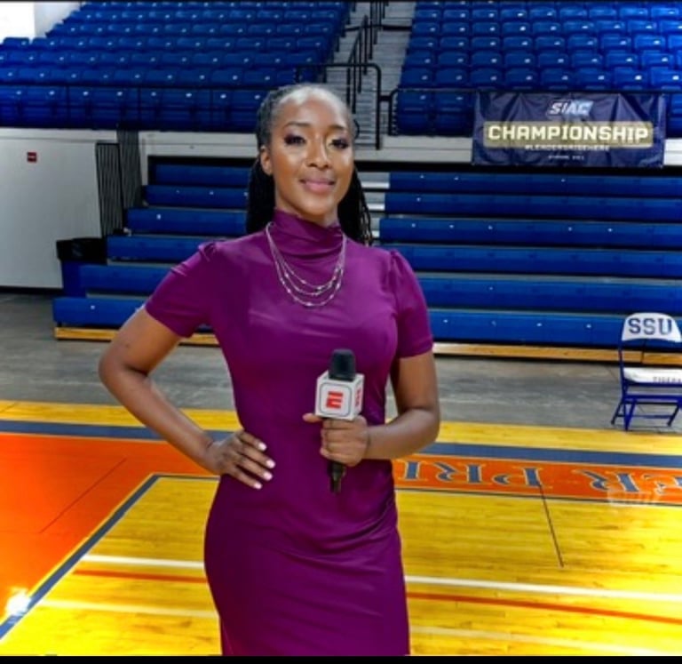 Woman in burgundy dress holding microphone on basketball court with blue bleachers behind her