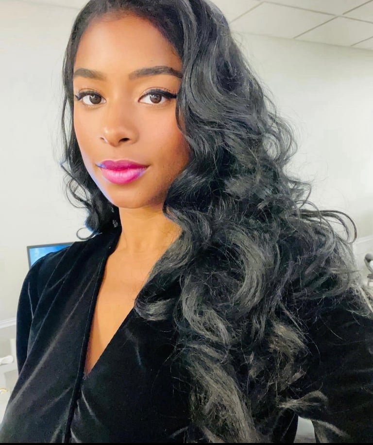 Woman with long black wavy hair and bright pink lipstick wearing a black velvet jacket, posing for a portrait indoors.