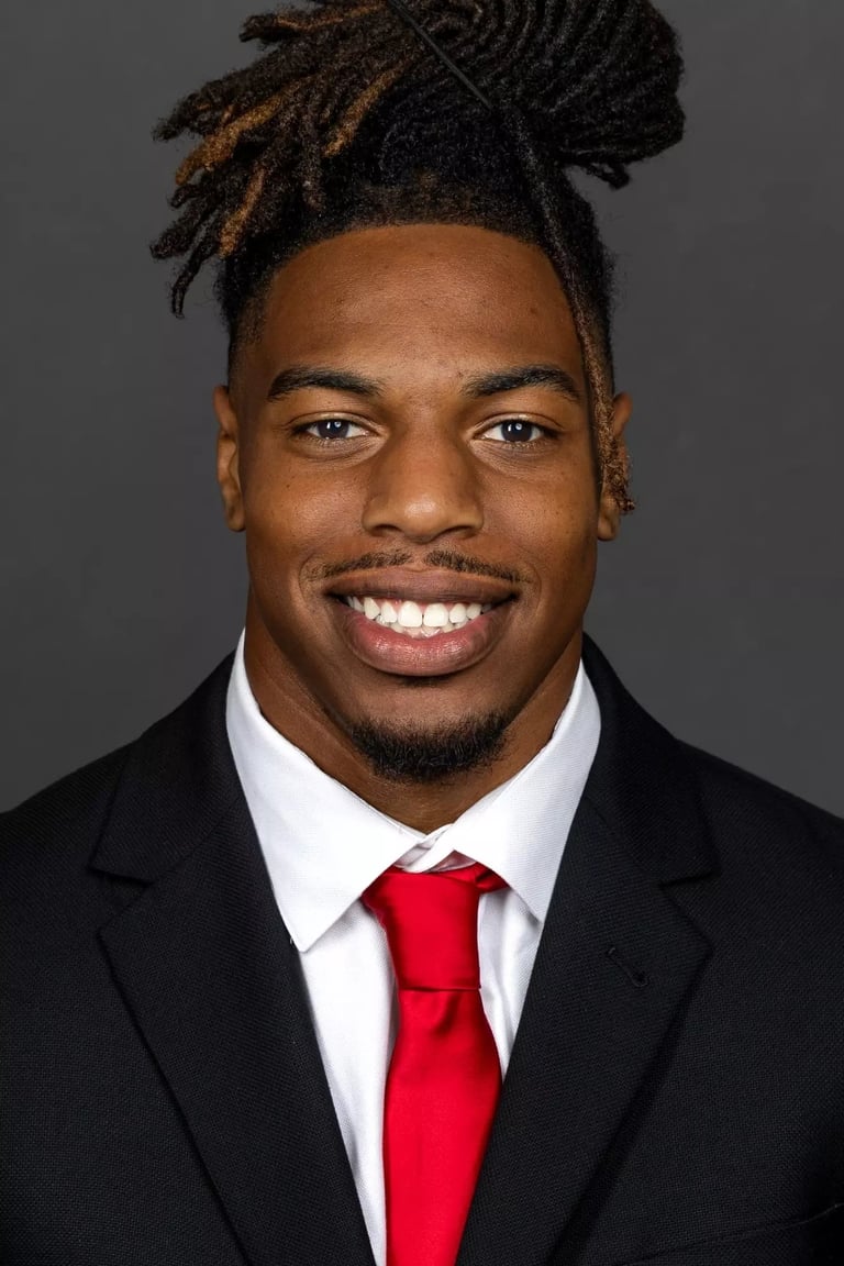 Professional headshot of a smiling man wearing a black suit, white dress shirt, and red tie against a gray background