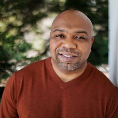 Smiling man with bald head wearing a rust-colored shirt, photographed outdoors with blurred green foliage in background