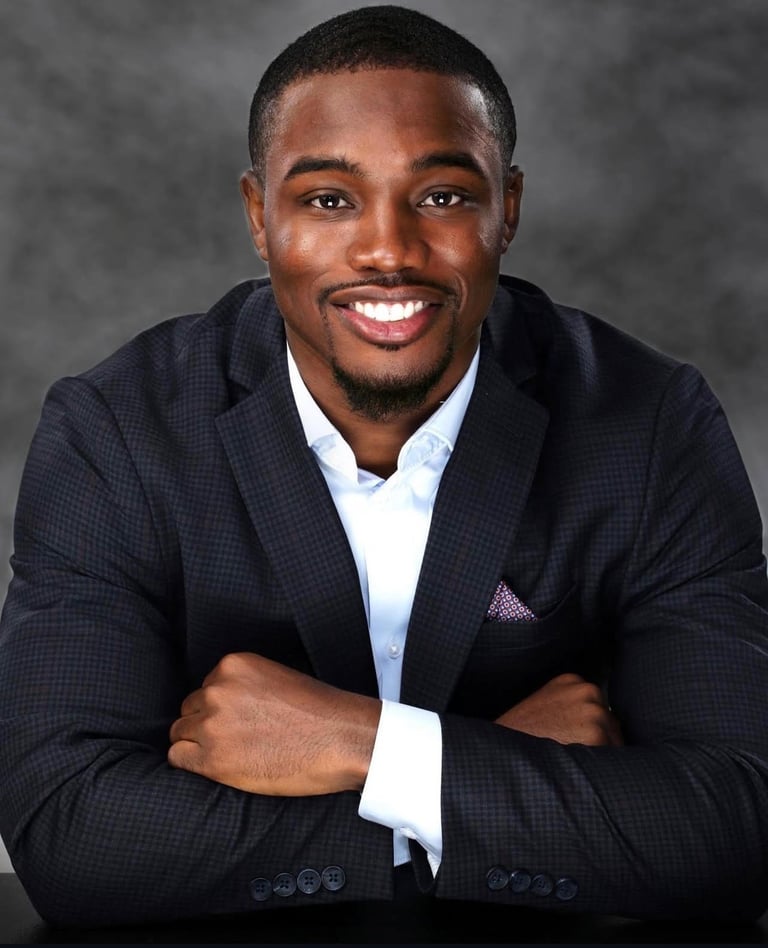 Professional headshot of a smiling man in a dark checkered blazer with arms crossed against a gray background
