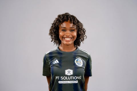 Smiling young woman with curly dark hair wearing a dark green PT Solutions Physical Therapy sports jersey against a light gray background